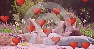 Animation of red heart balloons over happy diverse couple relaxing at picnic in sunny garden