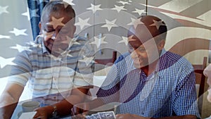Animation of waving flag of america over senior diverse friends playing cards at home