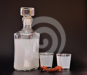 Anise liqueur, a large alcoholic drink in a glass bottle and two glasses on a black background, next to the stars of dried spices