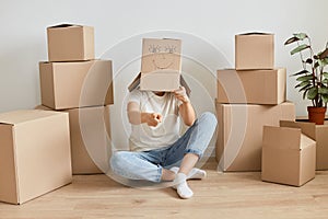 Anknown woman wearing white t shirt and jeans sitting on floor surrounded with cardboard boxes and putting on her head carton