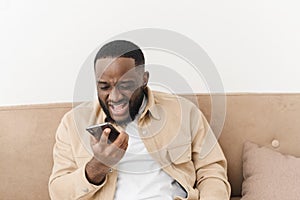 Annoyed african american man yelling at phone while sitting on sofa at home. Angry man looks at the phone while reading