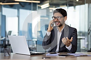 Annoyed and angry hispanic businessman talking on phone, man at workplace inside office with laptop