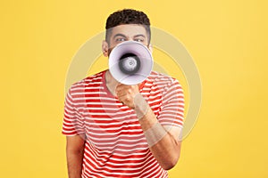 Annoyed angry man in striped t-shirt holding megaphone near mouth loudly speaking, screaming, making announcement, paying