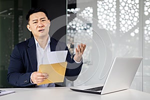 Annoyed asian businessman looking at camera and showing received letter with bad news, man working inside modern office