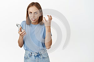 Annoyed blond girl holding her mobile phone, clench fist in anger, staring furious and irritated, bothered by someone