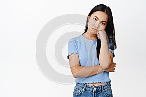 Annoyed or bored asian girl lean on hand, looking pissed off and offended at camera, standing in blue tshirt over white