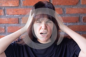 Annoyed brunette woman with bangs wearing a black t-shirt screaming due to conflict, showing all anger and anger on brick wall