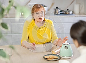 Annoyed elderly woman with cup of tea in her hands in kitchen listening to her friend photo