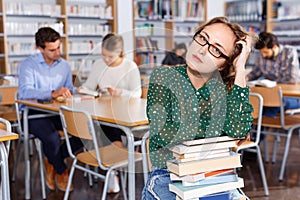 Annoyed girl sitting with pile of books in high school library on background of working fellow students