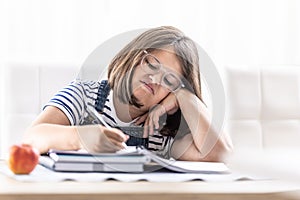 Annoyed schoolgirl with eyewear writes homework leaning against her hand