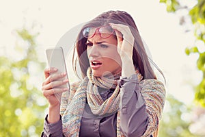 Annoyed upset woman in glasses looking at her smart phone with frustration while walking on a street