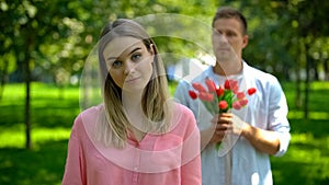 Annoyed young female looking camera, loving young man with flower on background