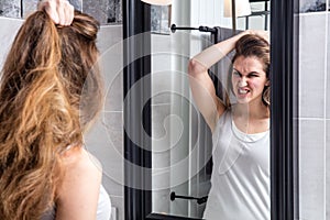 Annoyed young girl holding her damaged long hair in mirror