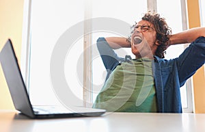 Annoyed young man with curly hair sitting at his desk with laptop feeling upset.