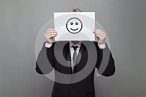 Anonymous businessman hiding his head behind happy smile emoticon on gray background