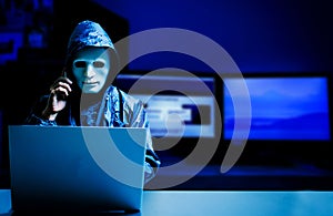 Anonymous computer hacker in white mask and hoodie.Obscured dark face using laptop computer for cyber attack and calling