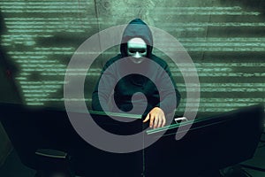 Anonymous hacker wearing face mask working on computer in dark room