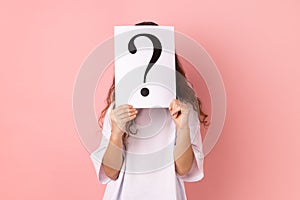 Anonymous little girl wearing white T-shirt hiding her face behind white paper with question mark.