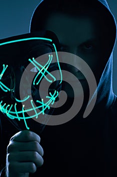 Anonymous man in black hoodie hiding his face behind a neon mask