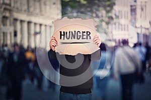 Anonymous man covers his head using a torn cardboard banner with hunger text message. Incognito person on a crowded street as