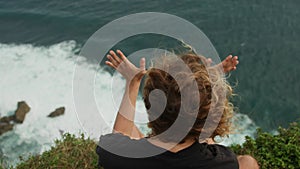 Anonymous man with long curly hair sitting on edge of ocean cliff moving hands as if controlling power of sea waves