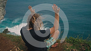Anonymous man with long curly hair sitting in lotus position on edge of ocean cliff moving hands as if controlling power