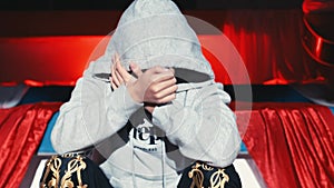 Anonymous person in hoodie covering face with hands, sitting on stage with red curtain backdrop