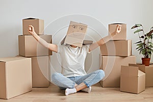 Anonymous woman wearing white t shirt and jeans sitting on floor, posing with cardboard box on her head, raising hands, rejoicing