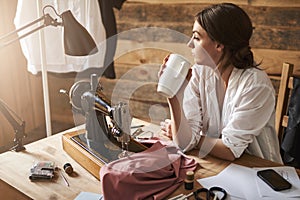 Another day in workshop came to end. Dreamy thoughtful female sewer looking aside while sitting near sewing machine