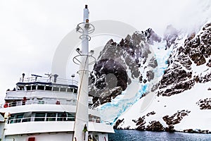 Antarctic cruise ship front view with mast and huge steep stone rock covered with glacier, close to Argentine islands, Antarctica