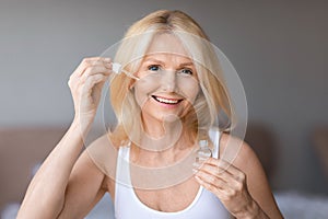 Anti-age cosmetics. Mature woman applying face serum with droplet on cheek, attractive smiling female moisturising skin