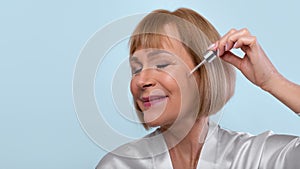 Anti-aging cosmetics. Beautiful mature woman applying regenerating facial serum onto her skin over blue background