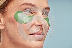 Anti aging procedures. Close up portrait of a beautiful adult lady using eye patches while standing against blue