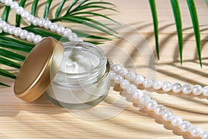 Anti-wrinkle moisturizing face cream in an open jar and pearl beads on a wooden desk. Green leaf casts a shadow on the glass