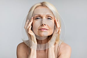 Antiage skincare. Senior woman touching skin near eyes and looking at camera, lady with flawless skin, grey background