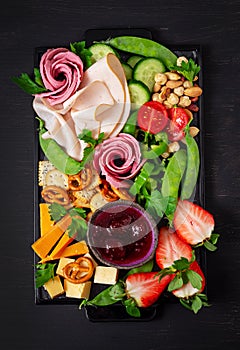 Antipasto platter cold sliced ham, salami, crackers, strawberries, vegetables and cheese platter on  board over dark background.