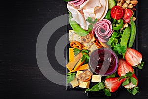 Antipasto platter cold sliced ham, salami, crackers, strawberries, vegetables and cheese platter on  board over dark background.