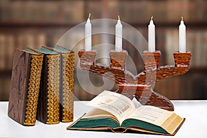 Antique books with an open book in front of an antique wooden candlestick on a bright table over abstract blurred library.