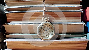 Antique gold pocket watch on chain hanging against background of stacks of books. Retro clock near old textbooks with