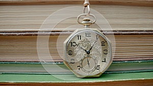 Antique gold pocket watch on chain hanging against background of stacks of books. Retro clock near old textbooks with