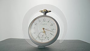 Antique grey pocket watch with a white dial and gold hands is spinning in bright light. Round mechanical vintage pocket