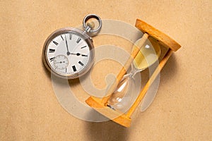 Antique pocket watch and glass hourglass on beige background. Round grey retro clock with black hands and white dial