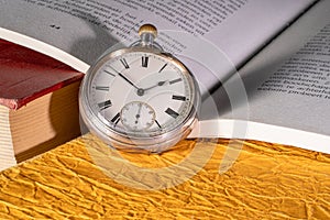 Antique silver pocket watch on background of an open book with text. Round vintage clock near old books with red and