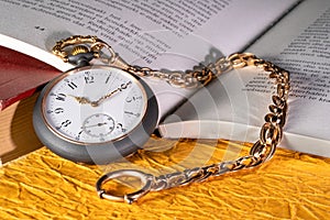 Antique silver pocket watch with gold chain lying on pages open book with text. Round grey vintage clock near old books