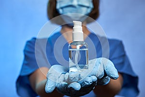 Antiseptic bottle in hands of medical worker wearing medical uniform, blue latex gloves and protective mask, COVID-19