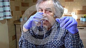 Antivirus protection. Covid-19. The senior man wearing in medical gloves drinks water, is sitting at a table in the kitchen.