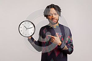 Anxious displeased man with dreadlocks and beard pointing finger on wall clock in his hand. I need more time