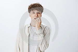 Anxious and insecure young teenage guy with curly hair in shirt biting fingernails looking unsure and nervous as