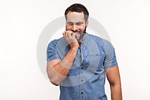 Anxious nervous man with beard in blue shirt biting nails on fingers looking at camera with terrified expression, confused and