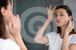 Anxious woman looking in mirror, touching forehead, confused about wrinkles